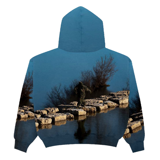 Back of a hoodie with a nature-themed design of a Sasquatch waking on rocks in between a lake on a blue background