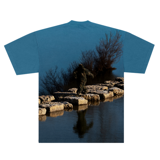 Back of a Blue shirt with a nature-themed design of a Sasquatch waking on rocks in between a lake on a blue background