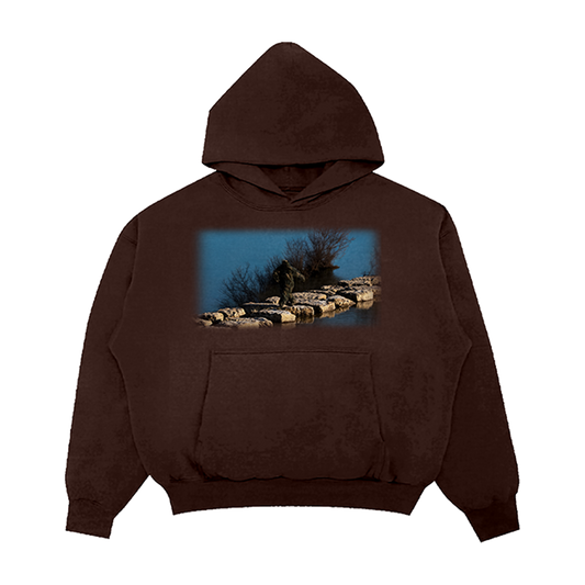 Brown hoodie with a scenic print on a nature-themed design of a Sasquatch waking on rocks in between a lake.