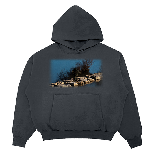 A dark charcoal gray pullover hoodie with a front kangaroo pocket and a rectangular graphic print on the chest depicting a person standing on rocks by a blue body of water.