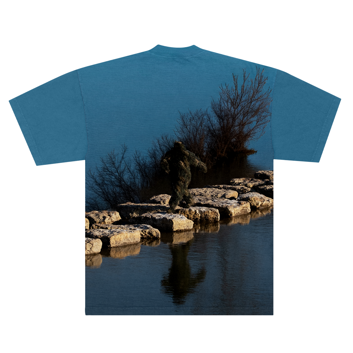 Back of a Blue shirt with a nature-themed design of a Sasquatch waking on rocks in between a lake on a blue background